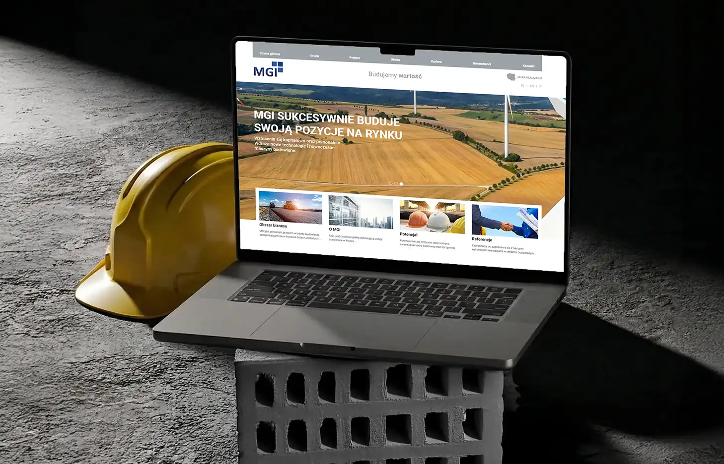 Strona internetowa dla firmy budowlanej wyświetlona na laptopie, w otoczeniu elementów budowlanych – kask i betonowy bloczek.