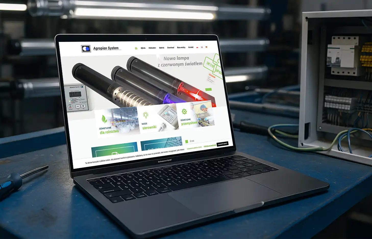 Laptop z otwartą stroną Agropian System na stole w hali produkcyjnej z oświetleniem LED i sterownikami przemysłowymi